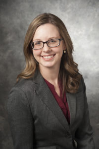 A professional headshot of a woman dressed in a gray jacket and maroon shirt. She is smiling and wearing blue glasses.